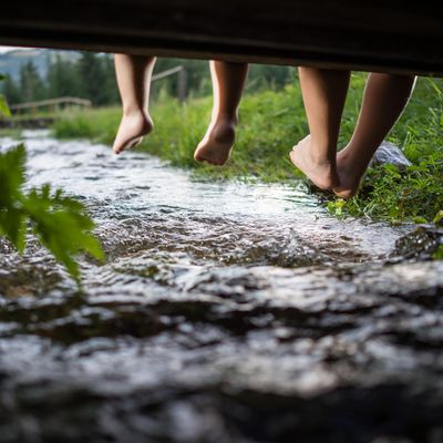 Aufnahme unter einer Brücke, unter der ein kleiner Bach fließt. Füße von zwei Personen hängen bei der Brücke herunter.