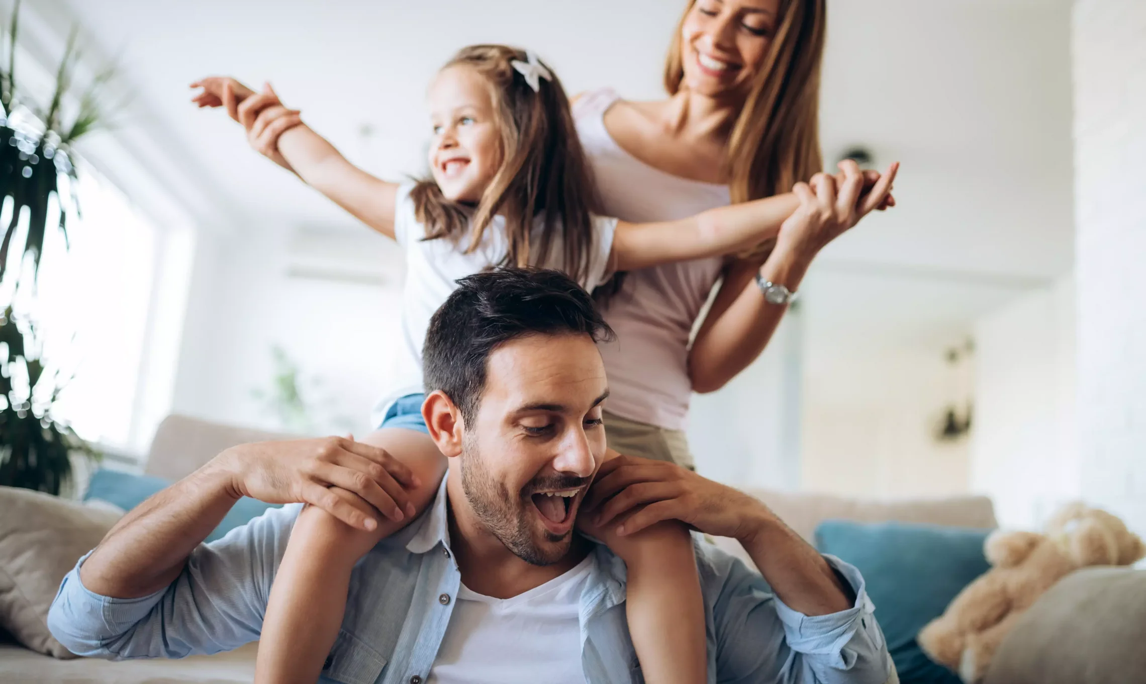 Ein Mädchen sitzt auf den Schultern von ihrem Vater. Hinter ihr steht ihre Mutter. Alle lachen und haben Spaß.